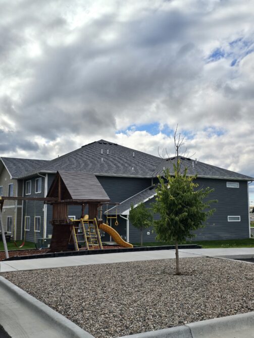 Playground with wooden structure, slide, and swings in front of gray residential building under cloudy sky