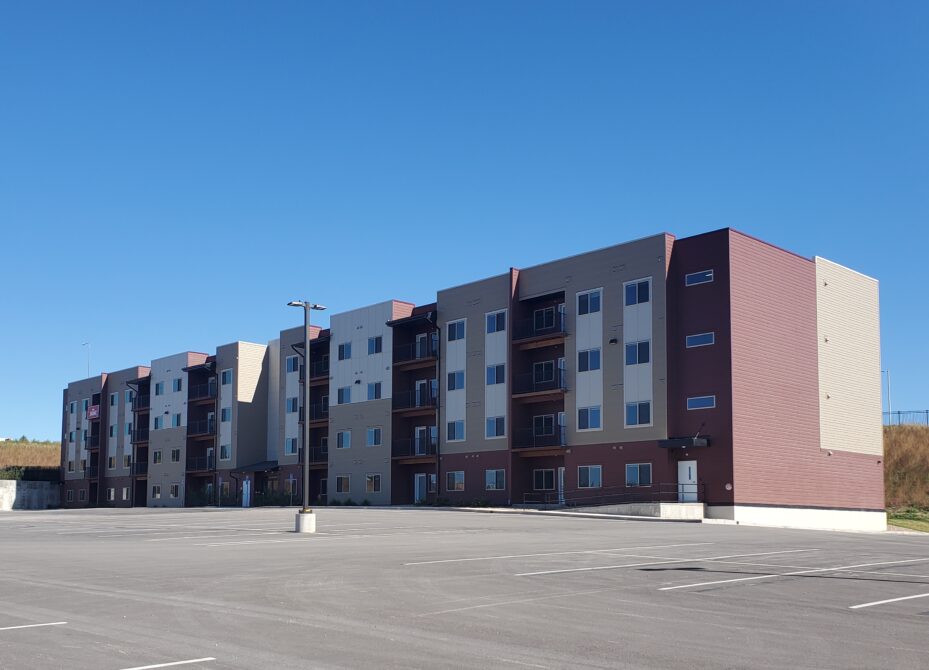 Four-story apartment building with multi-colored facade and empty parking lot. Clear blue sky.