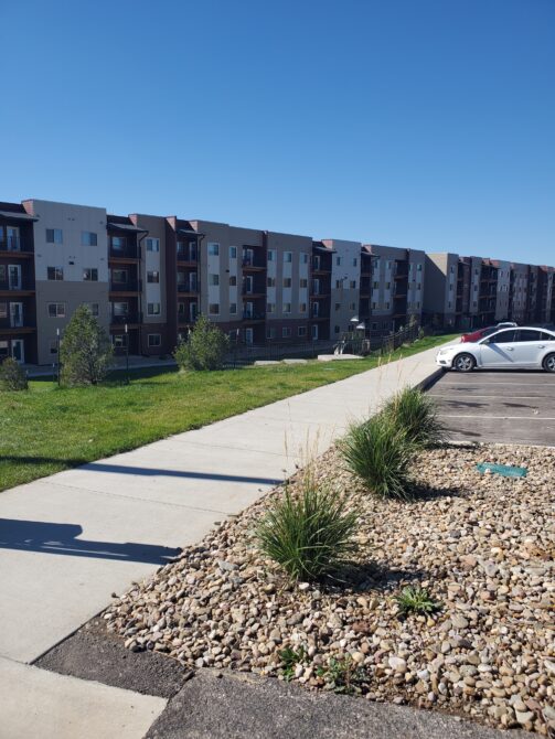 Residential apartment buildings with a parking lot and landscaped pathway under a clear blue sky