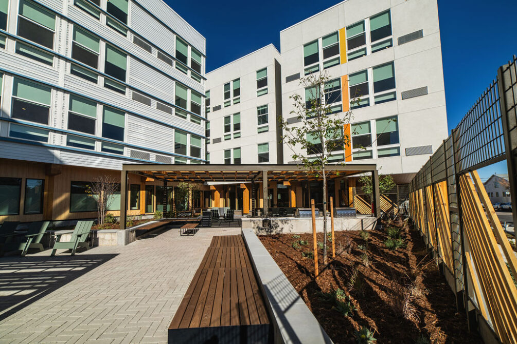Modern apartment courtyard with seating, young trees, and a pergola, surrounded by four-story buildings.