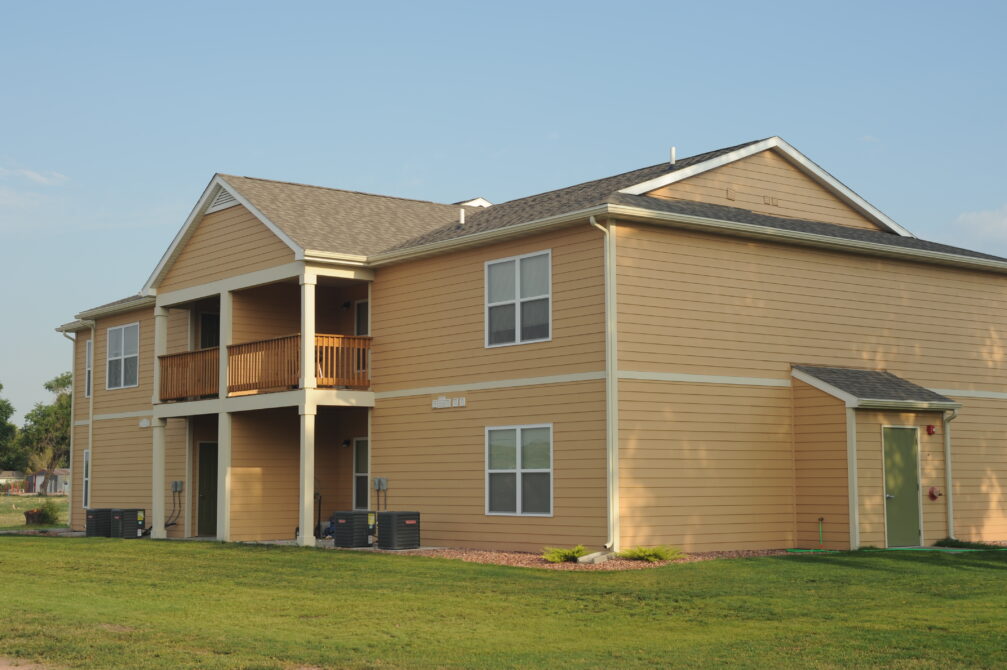 Two-story beige apartment building with wooden balcony and green door, surrounded by grassy lawn under clear sky.