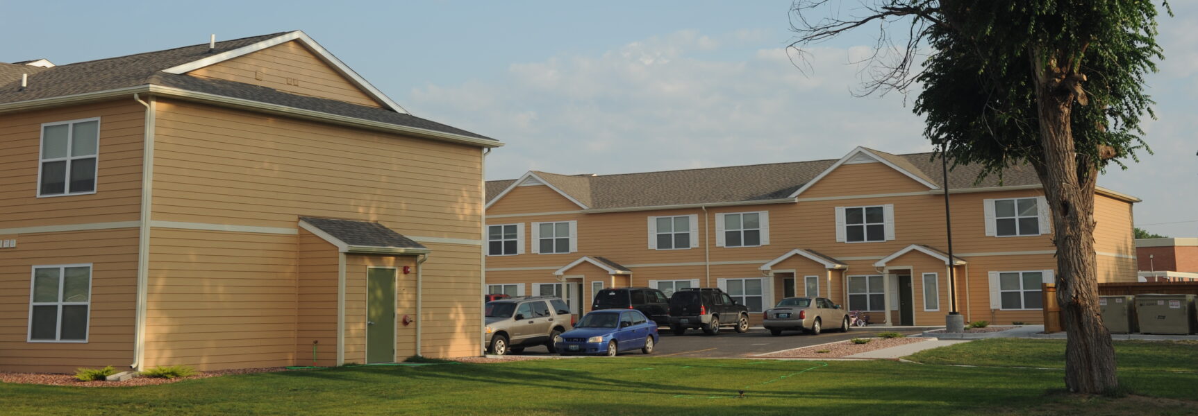 Tan two-story apartment buildings with parked cars and a tree