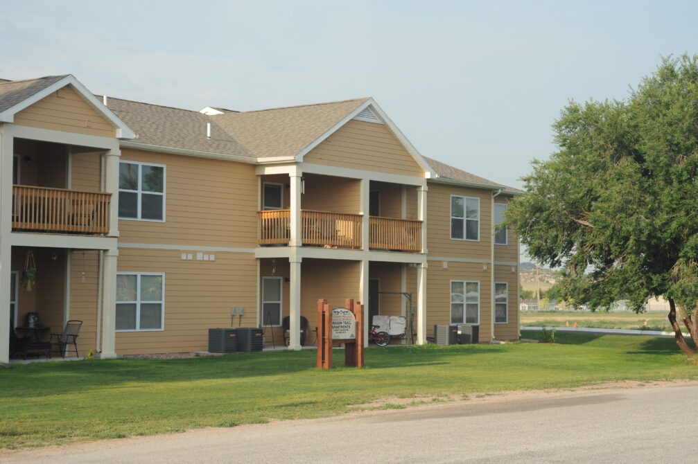 Two-story beige apartment building with wooden balconies, surrounded by green lawn. A wooden sign reads: “Brook View Apartments. 1885 East 8th Street. Leases Available.”