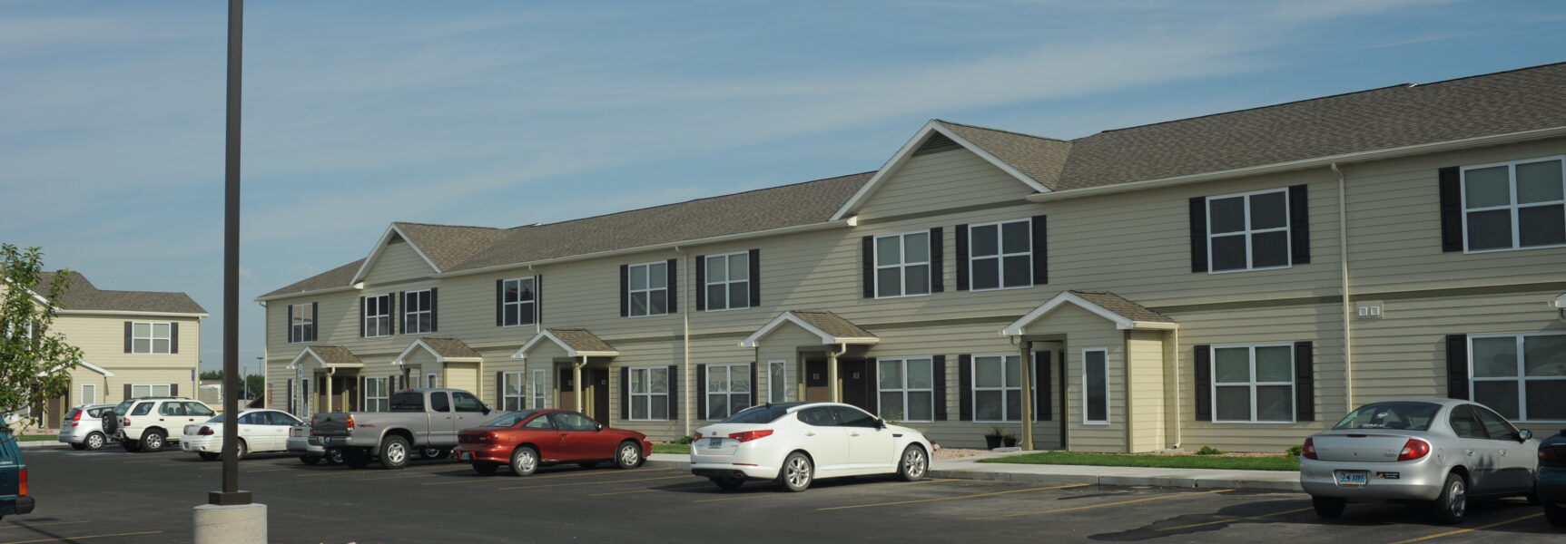 Two-story beige apartment complex with multiple entrances, parked cars in front, and clear blue sky above.