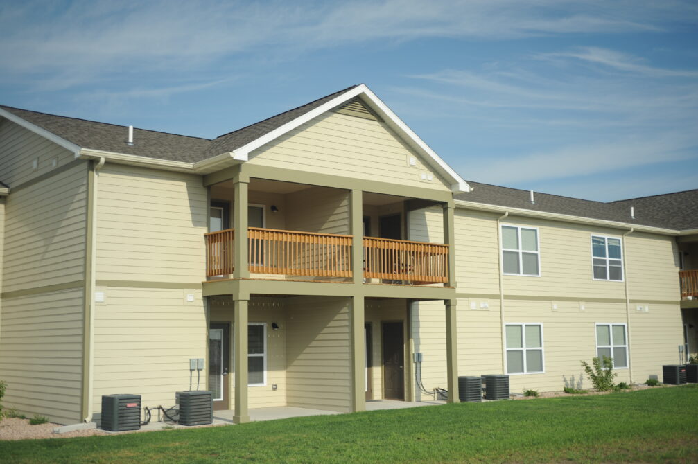 Two-story beige apartment building with wooden balcony, green lawn, and clear sky.