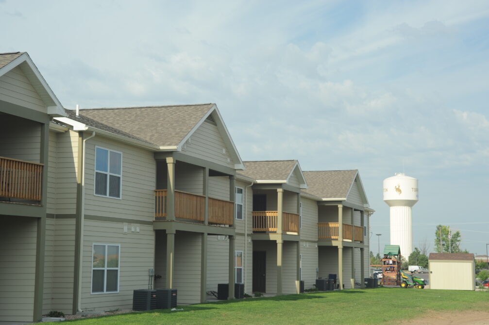 Row of tan apartment buildings with balconies, adjacent water tower labeled "Torrington" against a cloudy sky