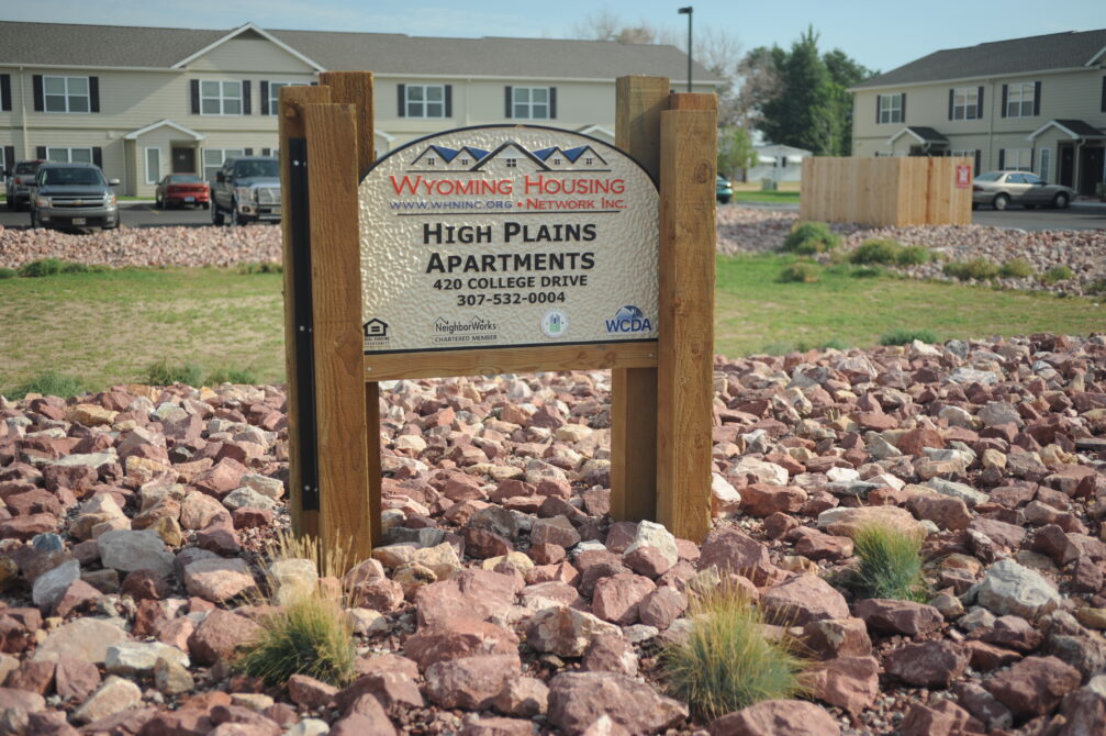 Sign for Wyoming Housing Network Inc, High Plains Apartments, 420 College Drive, 307-532-0004, surrounded by stones, buildings nearby