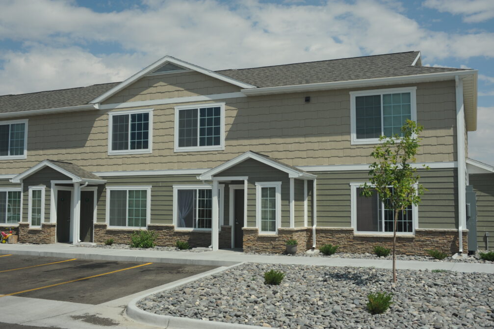 Two-story beige townhouse with white trim, stone accents, and a small young tree in the front.
