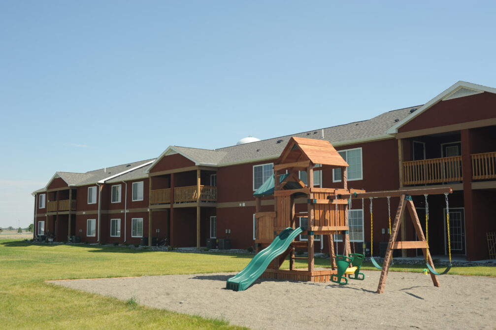 Two-story red apartment building with balconies, adjacent playground featuring swings and slides, clear blue sky overhead