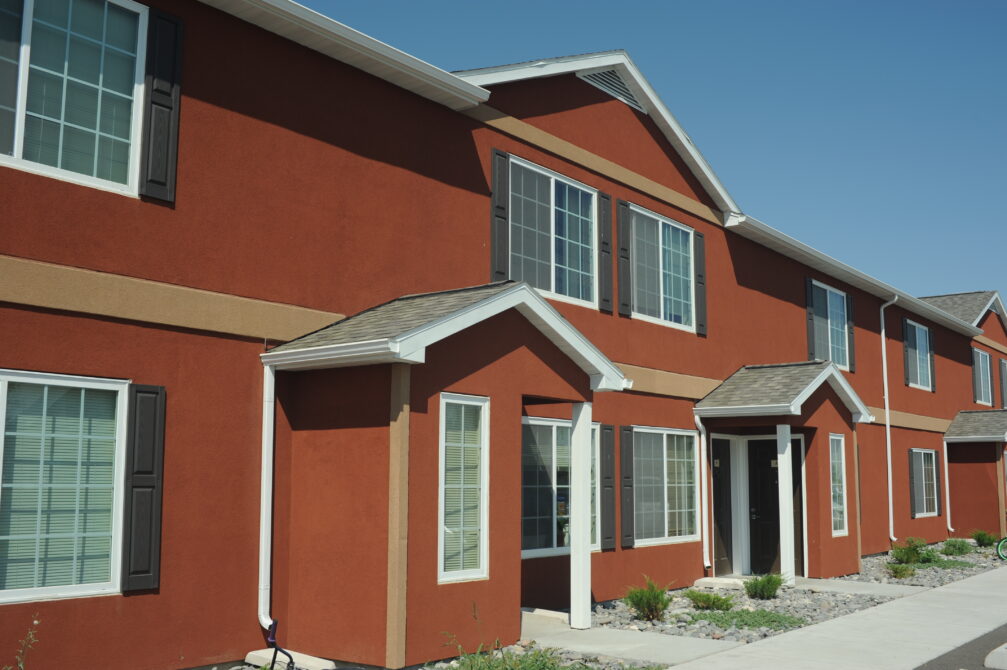 Red two-story apartment building with white trim, large windows, and small garden beds, under a clear blue sky