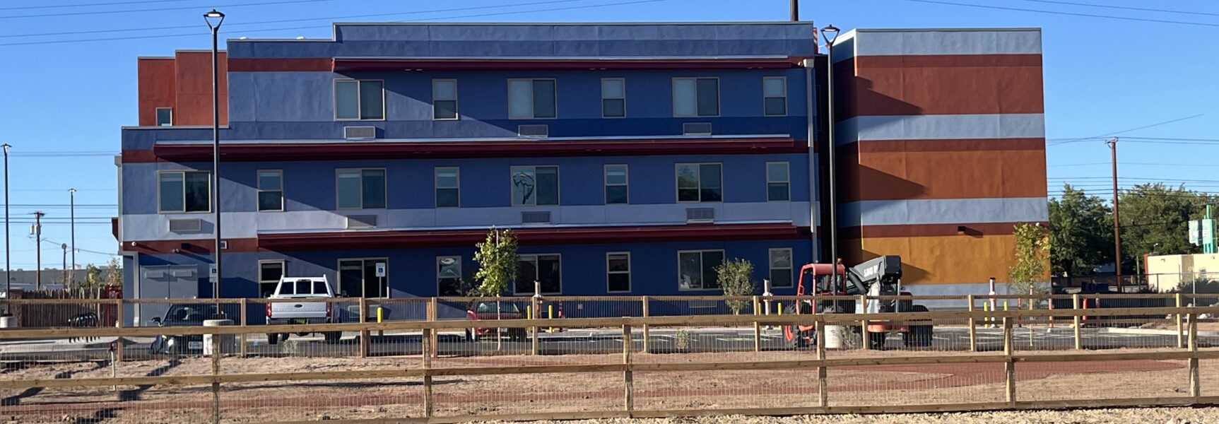 Colorful three-story building with parking lot, fenced foreground, and clear blue sky.