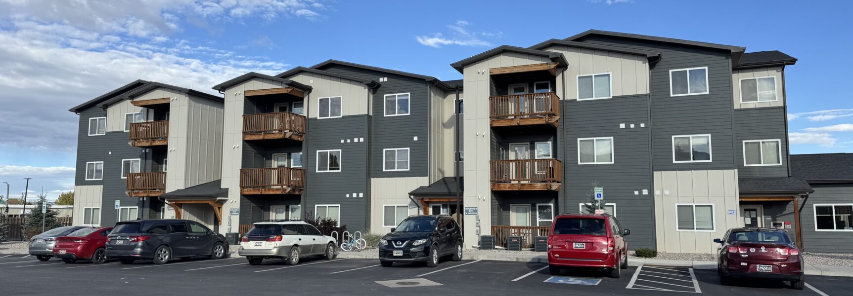 Three-story apartment building with wooden balconies, gray siding, parking lot with cars, and blue sky above