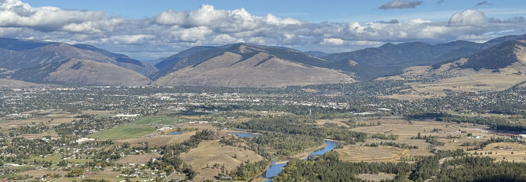 Aerial view of a sprawling valley town with mountains, winding river, and scattered clouds.