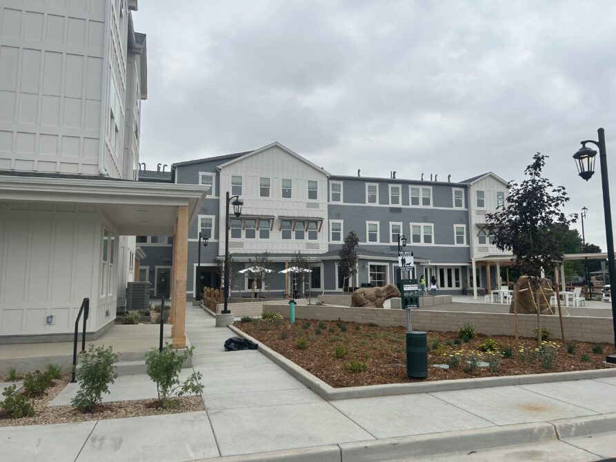 Modern three-story apartment building with a landscaped front yard and patio area under a cloudy sky.