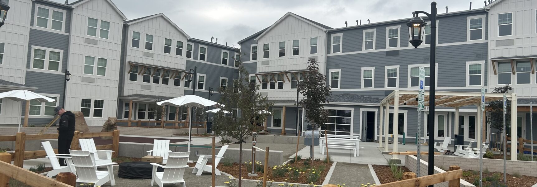 Modern apartment complex with outdoor seating area, white chairs, umbrellas, and landscaping under a cloudy sky
