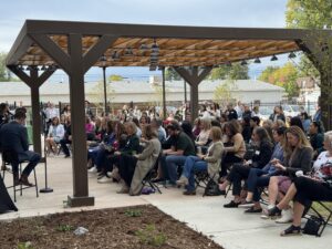 Audience attentively listening to speakers under a wooden pergola outdoors.