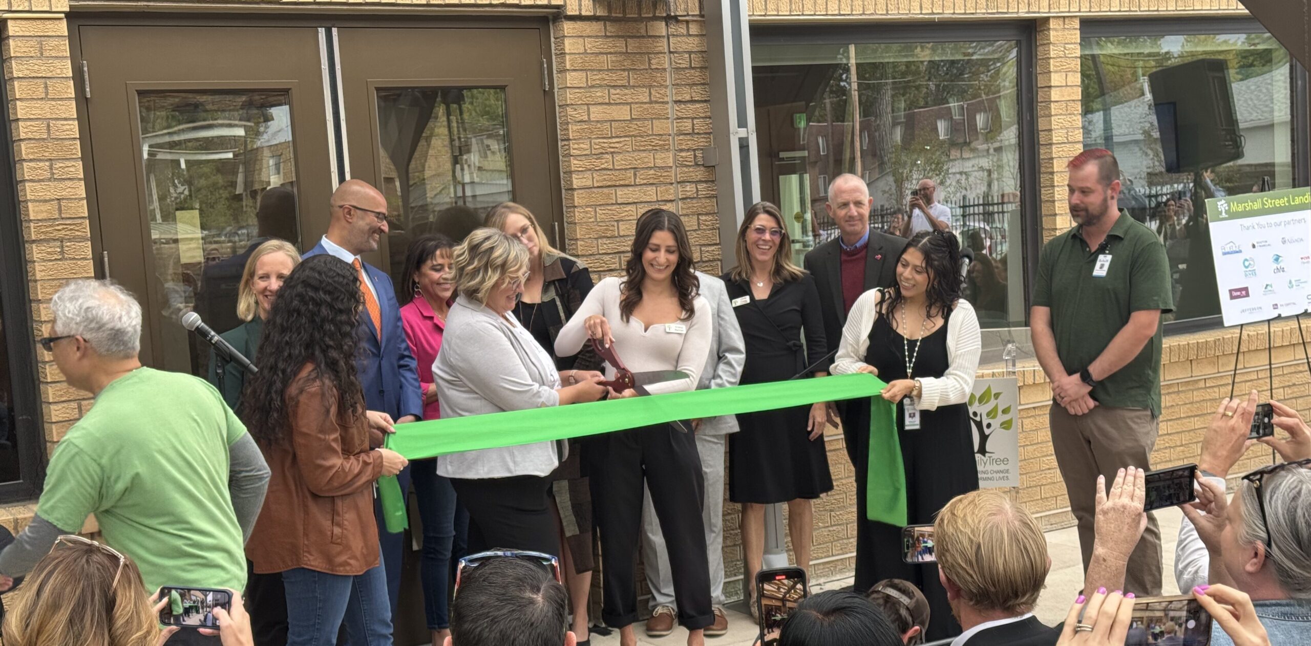 A group of people participate in a green ribbon-cutting ceremony outside a building, with FamilyTree signage visible.