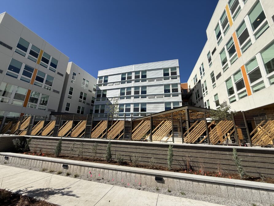 Modern apartment complex with light gray facade, large windows, and wooden accents under a clear blue sky.
