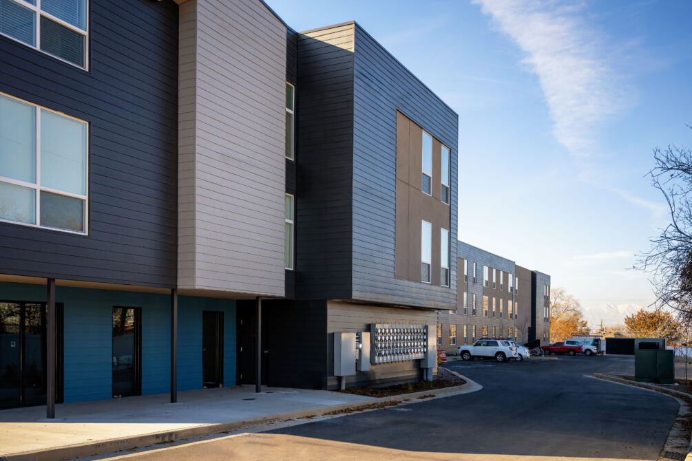 Modern apartment building exterior with large windows, overhanging facade, and parked cars in sunny setting.