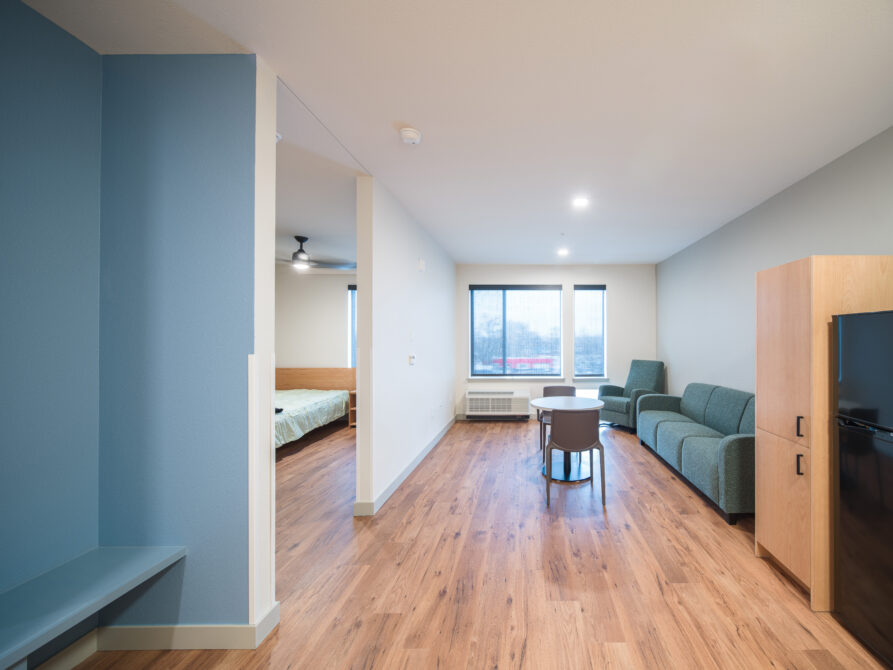 Modern living area with wooden floors, green sofa, round table, visible bedroom.