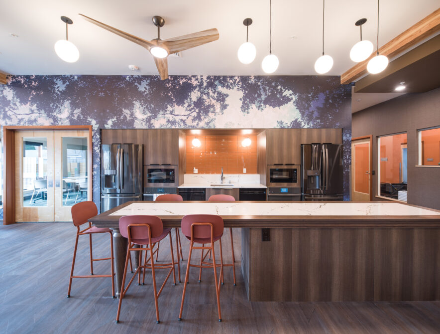 Modern kitchen interior with large island, hanging lights, and abstract wall design. Orange high chairs at the counter.