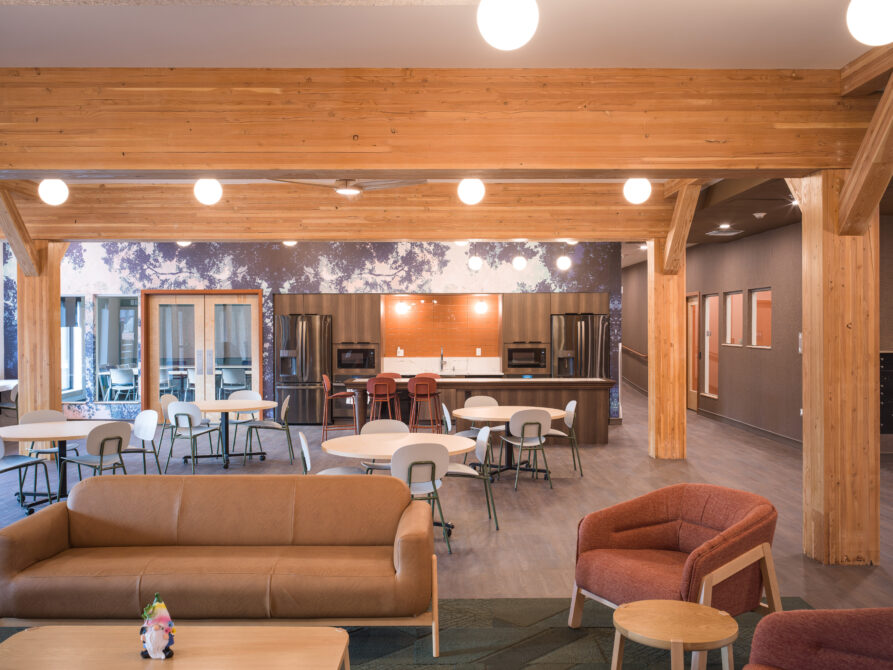 Spacious modern communal area with wooden beams, seating, tables, and a large kitchen in the background.