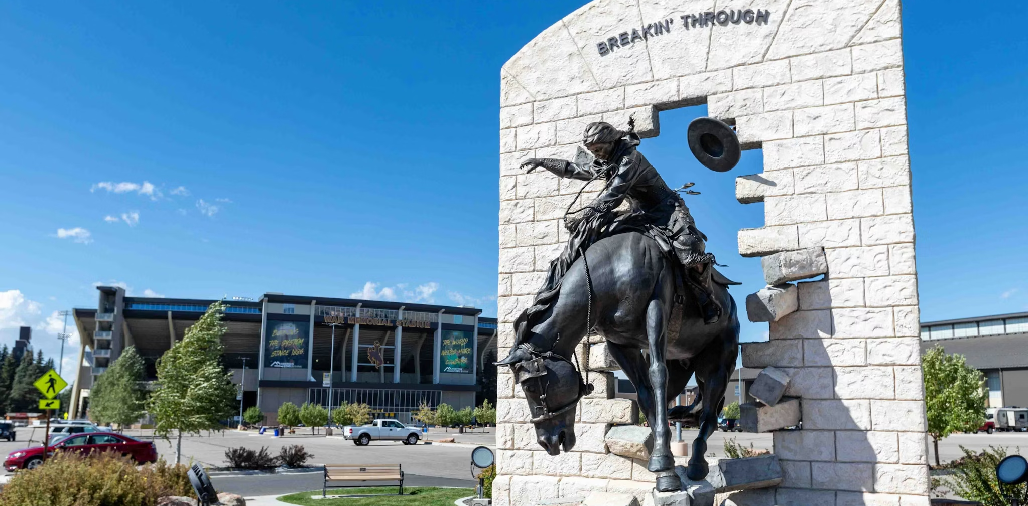 Statue of a rider and horse breaking through a stone wall, titled "Breakin' Through," with a stadium in the background.